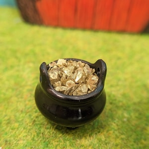 Op de afbeelding: Een kleine zwarte ketel gevuld met gouden glitter, een miniatuur rekwisiet voor een feeëntuin of poppenhuis.