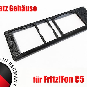 May include: Black replacement housing for a Fritz!Fon C5 phone. The housing has a textured surface and two rectangular cutouts. The text "Ersatz Gehäuse" and "für Fritz!Fon C5" is printed on the image.