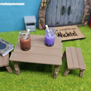 Könnte beinhalten: Zwei Miniatur-Glasgefäße mit Strohhalmen, eines gefüllt mit einer braunen Flüssigkeit und das andere mit einer violetten Flüssigkeit, stehen auf einem braunen Holztisch mit zwei passenden Bänken. Der Tisch ist im Freien auf einer grünen Grasfläche aufgebaut.