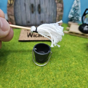 Może przedstawiać: Miniaturowy biały mop z drewnianym trzonkiem i czarnym wiadrem. Mop jest trzymany przez rękę, a wiadro stoi na zielonej powierzchni.