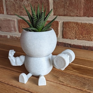 Può includere: Vaso da fiori in ceramica bianca a forma di persona che solleva pesi, con una piccola pianta grassa verde che cresce al suo interno.
