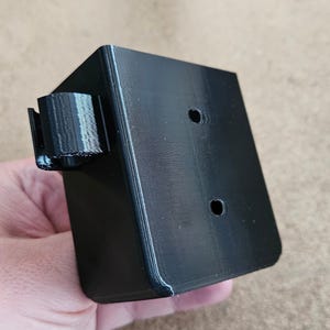 Può includere: Supporto a parete in plastica nera con due fori e una clip sul lato.