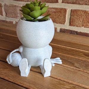 Könnte beinhalten: Ein weißer Keramik-Pflanztopf in Form einer sitzenden Figur mit einer kleinen Sukkulentenpflanze darin. Der Topf hat eine strukturierte Oberfläche und die Figur hat einen runden Kopf und zwei Arme.