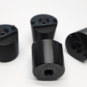 Könnte beinhalten: Vier schwarze, zylindrische Objekte mit strukturierter Oberfläche und einem Loch in der Mitte. Die Objekte sind auf einer weißen Oberfläche angeordnet. Die Objekte scheinen 3D-gedruckt zu sein.