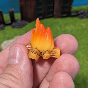 Puede incluir: Una figurita de fuego de campamento naranja y amarilla en miniatura con troncos marrones. La figurita está hecha de plástico y mide aproximadamente 5 cm de alto.