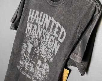 May include: Black t-shirt with a vintage-style graphic of the Haunted Mansion logo and characters. The text reads "Haunted Mansion" and "We've been dying to have you".