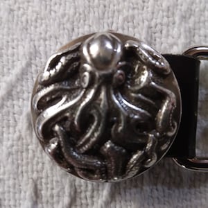 May include: A silver-colored belt buckle with a detailed octopus design. The round buckle is attached to a black leather strap and a silver-colored metal loop. The octopus is the focal point of the buckle.