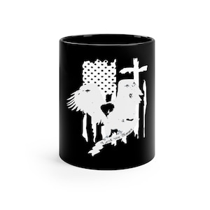 May include: Black ceramic mug featuring a white design of an eagle with outstretched wings, a cross, and an American flag. The text "Lest We Forget" is displayed on a banner beneath the eagle.
