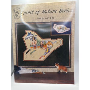 May include: A cross-stitch pattern titled "Spirit of Nature Series: Horse and Fox" by Designer Mike Vickery. The artwork features a horse and fox design with Native American imagery. A small fox figurine and a decorative feather are in the foreground.