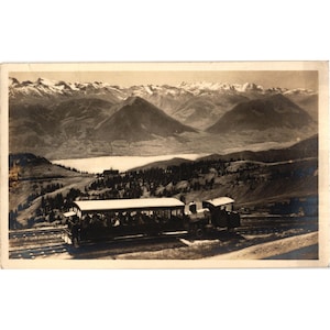 May include: Vintage black and white postcard featuring a train carrying passengers on tracks. The train travels through a mountainous landscape with snow-capped peaks, a lake, and lush greenery. The postcard has a sepia tone.