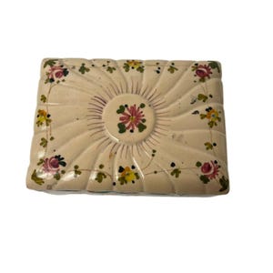 May include: Cream-colored rectangular ceramic box with a floral design. The box features a central sunburst pattern and is adorned with pink, yellow, and green floral accents. The edges are slightly raised, adding dimension to the piece.