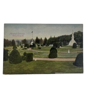 May include: Vintage postcard featuring the National Cemetery in Gettysburg, PA. The image shows a well-manicured park with monuments, trees, and trimmed bushes. The sky is overcast, and the postcard has a slightly faded appearance, suggesting its age.