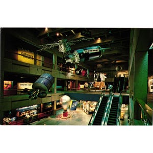 May include: Vintage postcard featuring a museum interior with space-themed exhibits. The image showcases a large model of a space station, a lunar module, and other spacecraft suspended from the ceiling. The museum has escalators and multiple levels.