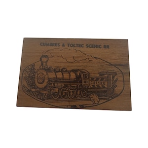 May include: A wooden plaque featuring an engraved image of a classic steam train. The text "Cumbres & Toltec Scenic RR" is above the train illustration. The plaque has a natural wood grain finish.