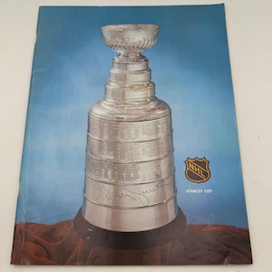 May include: A silver Stanley Cup trophy with a detailed design, set against a blue background. The NHL logo and the words "STANLEY CUP" are visible. The trophy sits on a brown surface.