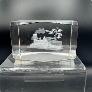 May include: A clear, rectangular crystal block featuring a 3D etched image of a steam train, a tree, and smoke. The train is on tracks, with detailed etching within the glass. The block is displayed on a clear stand.