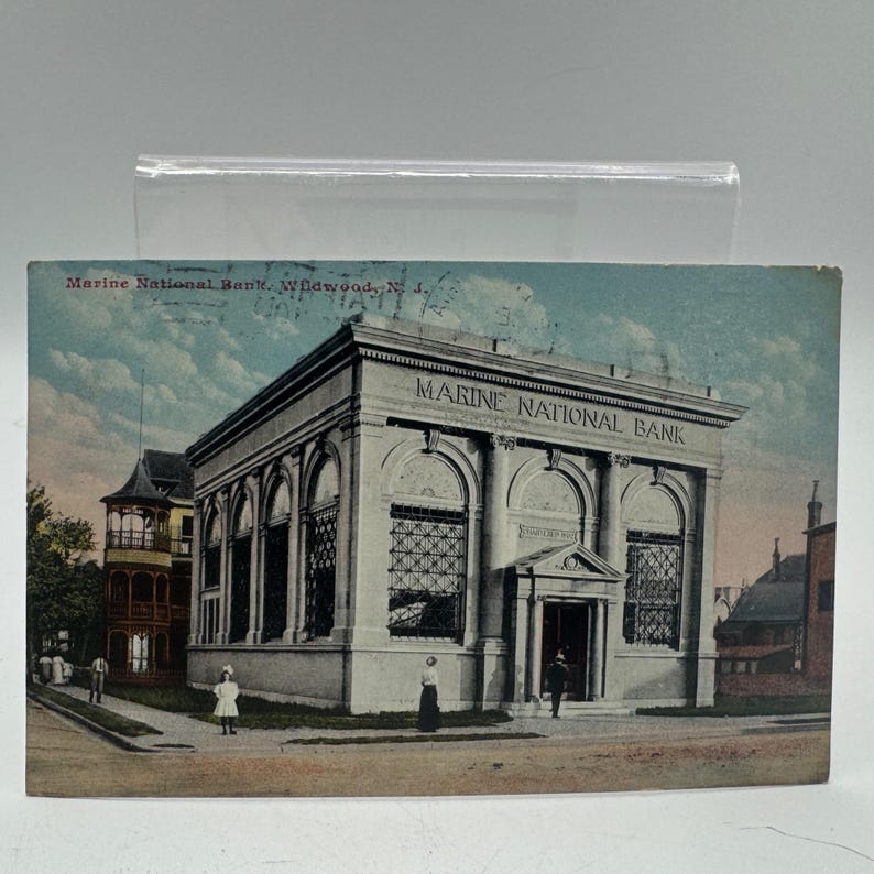 K&ouml;nnte beinhalten: Vintage Postkarte mit der Marine National Bank in Wildwood, N.J. Das Geb&auml;ude ist hellbeige mit gro&szlig;en Bogenfenstern und S&auml;ulen. Der Text "MARINE NATIONAL BANK" steht oben. Die Postkarte hat einen leichten Sepia-Ton.