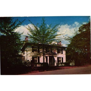 May include: Vintage postcard featuring a two-story white house with black shutters and a porch. The house is surrounded by lush green trees under a blue sky with fluffy white clouds. A road is in the foreground.