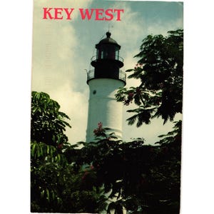 May include: A vintage postcard featuring the text "KEY WEST" in red letters. The image showcases a white lighthouse with a black top, surrounded by lush green foliage and a partly cloudy sky.