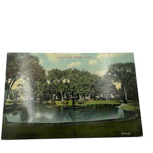 May include: A vintage postcard showing Chapin Park in Bangor, Maine. The postcard features a fountain in a pond surrounded by trees. The text "Chapin Park, Bangor, Maine." is printed in red at the top of the postcard.