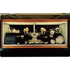 May include: A collection of silver serving pieces displayed in a wooden case, including a large centerpiece bowl, candelabras with shades, a pitcher, and various plates. A black plaque with white text titled U.S.S. NORTH CAROLINA SILVER SERVICE is visible.