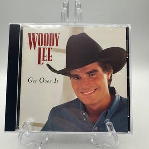 Puede incluir: Una caja de CD con un hombre sonriente con un sombrero de vaquero negro y una camisa de mezclilla. La portada dice "WOODY LEE" en letras rojas sobre el título "Get Over It."