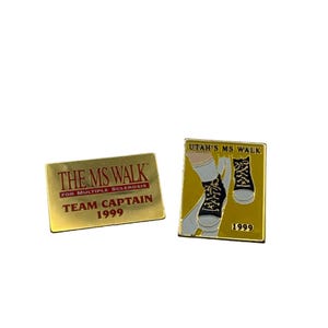 Puede incluir: Dos pines de oro con el texto "The MS Walk for Multiple Sclerosis Team Captain 1999" y "Utah's MS Walk 1999". El segundo pin presenta una imagen de dibujos animados de las piernas de una persona con zapatillas.