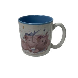 Op de afbeelding: Witte keramische mok met een blauwe binnenkant en een witte handgreep. De mok heeft een illustratie van drie roze varkens met haarbanden en strikken. Vlinders zijn verspreid rond de varkens.