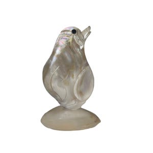 Op de afbeelding: Een gesneden, iriserend vogelbeeldje. De sculptuur is gemaakt van een parelmoerachtig materiaal, met een gladde, gepolijste afwerking. De vogel heeft een afgerond lichaam, een kleine snavel en een cirkelvormige basis. Het beeldje staat tegen een witte achtergrond.