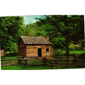 Op de afbeelding: Een vintage ansichtkaart met een rustieke blokhut met een houten hek in een groen landschap. De hut heeft een verweerd houten dak en een kleine schoorsteen. De scène is omgeven door bomen en een blauwe lucht.