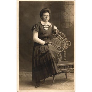Puede incluir: Una fotografía antigua en tono sepia que muestra a una persona con un vestido oscuro de cuello alto, mangas largas blancas y guantes oscuros. La persona está sentada en una silla de metal ornamentada con patrones de volutas.