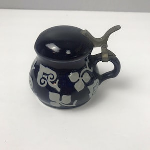 May include: A small, dark blue ceramic stein with a matching lid and a metal hinge. The stein features a white floral pattern and a handle. The lid is round and the handle is curved. This decorative stein is a unique addition to any collection.