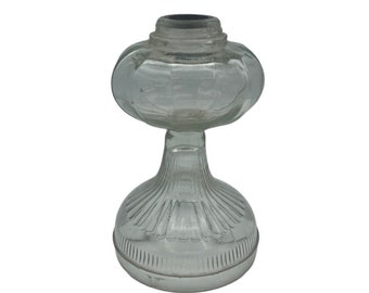 May include: A clear glass oil lamp with a round, ribbed base and a round, fluted top. The lamp has a black metal rim around the top.