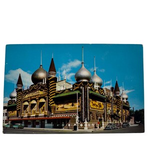 Könnte beinhalten: Eine Vintage-Postkarte mit einem farbenfrohen Gebäude in einem einzigartigen architektonischen Stil. Das Gebäude hat mehrere Türme mit zwiebelförmigen Kuppeln und ist mit Streifen und Wandgemälden verziert. Der Text "Mitchell Corn Palace" ist auf der Vorderseite des Gebäudes zu sehen.