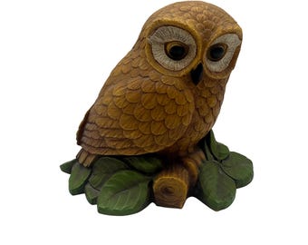 May include: A detailed brown owl figurine with large, dark eyes and textured feathers. The owl is perched on a small, green-leafed branch. The figurine is a decorative item.