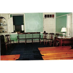 May include: A room with light green walls, a dark blue floor, and several wooden chairs and benches. A long wooden bench with spindle backrests sits against the wall, accompanied by a wooden table with a white lamp and a white door.
