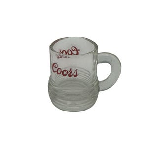 以下が含まれることがあります： 取っ手付きの小さな透明なガラスマグ。マグカップの下半分にはリブのデザインがあり、赤い文字で「Coors」と書かれています。マグカップは少量の液体を入れるように設計されています。