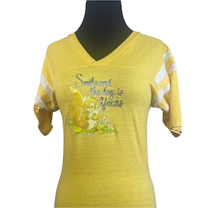 May include: Yellow short-sleeved t-shirt with white stripes on the sleeves. The shirt features a graphic of a squirrel holding a nut with the text "Smile and the day is yours".