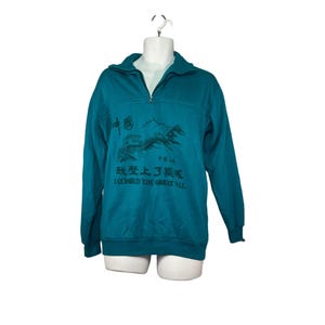 May include: Teal pullover sweatshirt with a quarter-zip neckline. The front features a graphic of the Great Wall of China with Chinese characters and the English text "I Climbed the Great Wall."