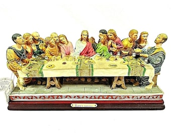 May include: A ceramic figurine depicting the Last Supper, featuring 13 figures seated around a table. The figures are painted in realistic detail, with various expressions and gestures. The table is covered in a white tablecloth with a green and gold pattern. The base of the figurine is made of wood and features a plaque with the text "Rudy's Collections".
