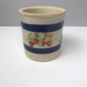 May include: A beige ceramic utensil holder with two navy blue stripes and a painted strawberry design. The cylindrical container has a slightly flared rim and is approximately 6 inches tall.