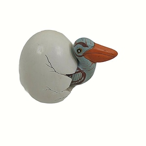 May include: A ceramic figurine of a blue and orange bird hatching from a white egg. The bird has a long orange beak and is partially out of the egg.