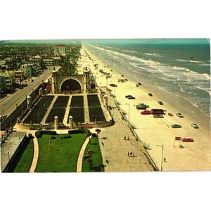 May include: Vintage postcard featuring an aerial view of a beach with a boardwalk, cars, and a large outdoor theater. The ocean is visible on the right, and a city with buildings and roads is on the left. The colors are faded, giving it a retro feel.