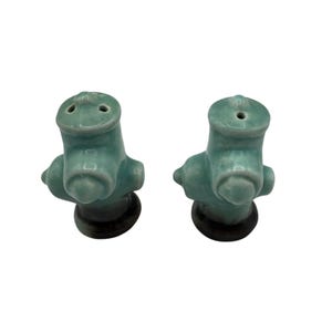 May include: A pair of ceramic salt and pepper shakers shaped like teal fire hydrants. Each shaker has a dark base and two holes on top for dispensing. The hydrants have a glossy finish and are set against a white background.