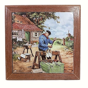 May include: Framed ceramic tile featuring a Dutch scene with a man carving wooden shoes. The artwork depicts a rural setting with a thatched-roof cottage, windmill, and tools. The frame is made of brown wood.