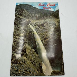 Puede incluir: Una postal vintage con una foto de Bear Creek Falls. La cascada cae por un acantilado rocoso, con árboles verdes exuberantes en el fondo. La postal tiene un cielo azul con nubes blancas.