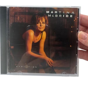 Può includere: Un'astuccio per CD con Martina McBride. La copertina dell'album mostra una donna con un top nero, seduta su una sedia. Il titolo dell'album è "Evolution". L'astuccio del CD è tenuto da una persona.
