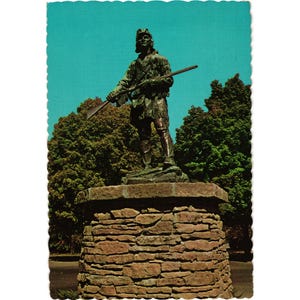 May include: Vintage postcard featuring a bronze statue of a frontiersman holding a rifle. The statue stands atop a stone pedestal against a backdrop of trees and a clear blue sky. The postcard has a scalloped edge.