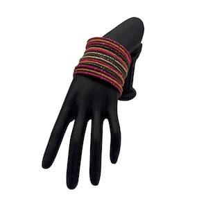 Puede incluir: Un conjunto de pulseras de cuentas en tonos rosa, dorado y marrón. Las pulseras se exhiben en un soporte negro con forma de mano. Las pulseras están hechas de pequeñas cuentas de colores. Fondo blanco.