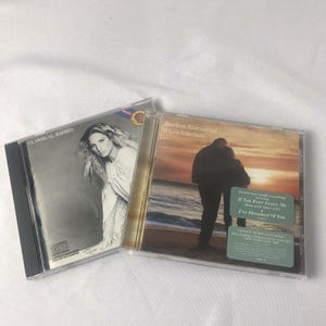 Könnte beinhalten: Zwei Barbra Streisand Musik-CDs in Kunststoffhüllen. Die linke Hülle zeigt ein Schwarz-Weiß-Porträt mit dem Titel Classical Barbra. Die rechte Hülle zeigt ein Paar an einem Strand bei Sonnenuntergang mit dem Titel A Love Like Ours und zwei Aufklebern.